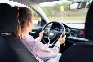 Woman driving car. Young driver. Testing vehicle in dealership or student training in school to get license. Hands on steering wheel. Behind back inside view of cockpit.